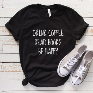 May include: Black t-shirt with white text that reads "Drink Coffee Read Books Be Happy". The shirt is laid flat on a white wooden surface with a pair of gray canvas shoes.
