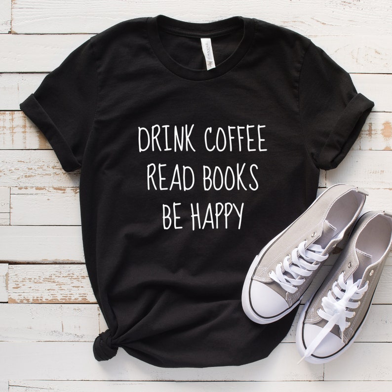 May include: Black t-shirt with white text that reads "Drink Coffee Read Books Be Happy". The shirt is laid flat on a white wooden surface with a pair of gray canvas shoes.