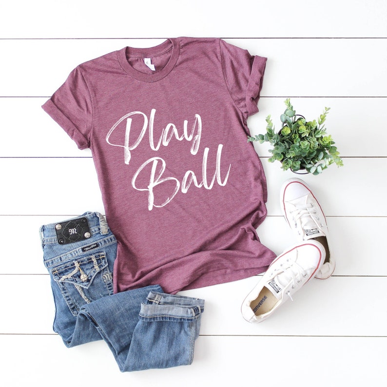 May include: A maroon t-shirt with the words "Play Ball" written in white on the front. The shirt is laying on a white wooden surface with a pair of white sneakers and a pair of blue jeans.