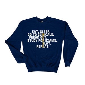 May include: Navy blue sweatshirt with the text "EAT. SLEEP. GO TO CLINICALS. FREAK OUT. STUDY FOR EXAMS. SLAY. REPEAT." in white. The word "NURSE" is spelled out vertically in yellow.