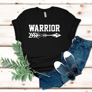 Puede incluir: Camiseta negra con un dise&ntilde;o de flecha y cinta blanca. El texto "WARRIOR" est&aacute; por encima de la flecha.