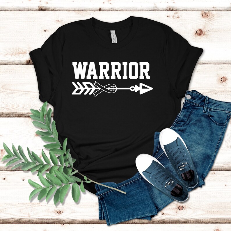 Puede incluir: Camiseta negra con un dise&ntilde;o de flecha y cinta blanca. El texto "WARRIOR" est&aacute; por encima de la flecha.