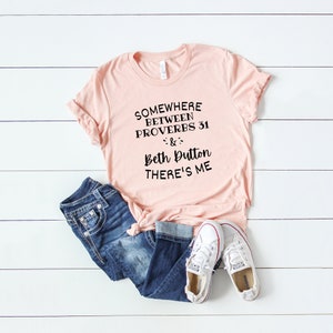 May include: A light pink short-sleeve t-shirt with black text that reads SOMEWHERE BETWEEN PROVERBS 31 & BETH DUTTON THERE'S ME, paired with dark blue denim jeans and white canvas sneakers with red and blue trim on the rubber soles.