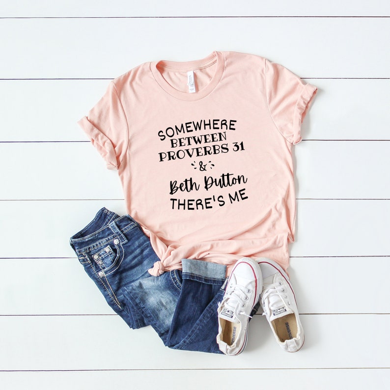 May include: A light pink short-sleeve t-shirt with black text that reads SOMEWHERE BETWEEN PROVERBS 31 & BETH DUTTON THERE'S ME, paired with dark blue denim jeans and white canvas sneakers with red and blue trim on the rubber soles.