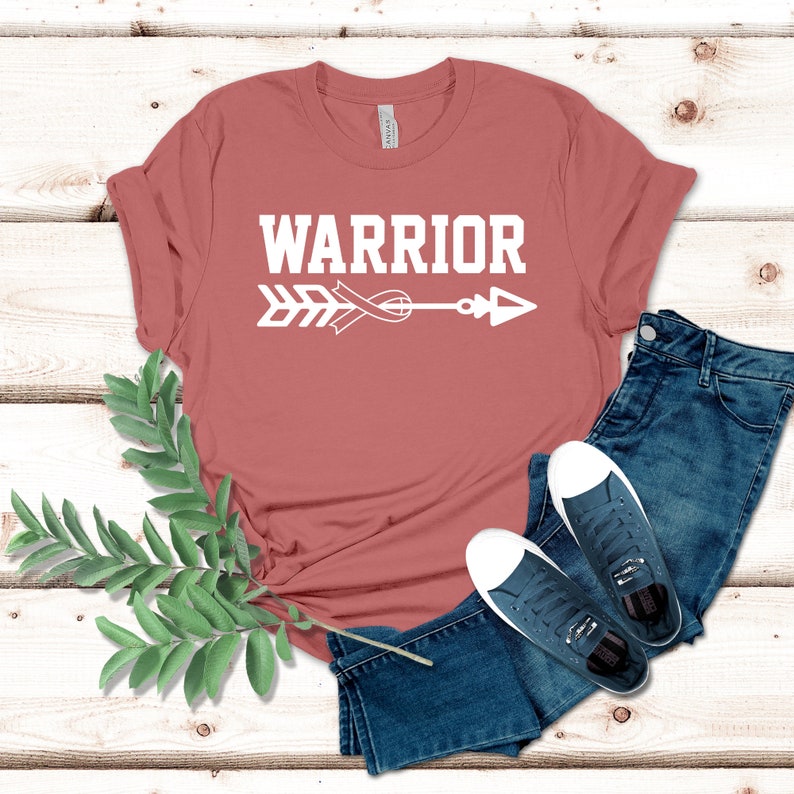 Puede incluir: Una camiseta de color &oacute;xido con la palabra "WARRIOR" impresa en blanco, junto con un dise&ntilde;o de flecha y cinta blancos.