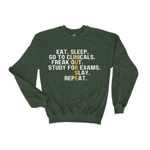 May include: Green sweatshirt with the text "EAT. SLEEP. GO TO CLINICALS. FREAK OUT. STUDY FOR EXAMS. SLAY. REPEAT." in white. The word "NURSE" is in yellow and vertical.