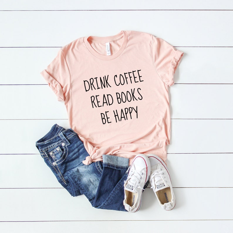 May include: A pink t-shirt with the text "Drink Coffee Read Books Be Happy" printed on it. The shirt is laid out on a white wooden surface with a pair of blue jeans and white sneakers.