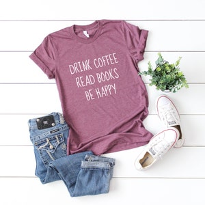 May include: A maroon t-shirt with the text "Drink Coffee Read Books Be Happy" in white lettering. The shirt is laid flat on a white wooden surface with a pair of blue jeans and white sneakers.