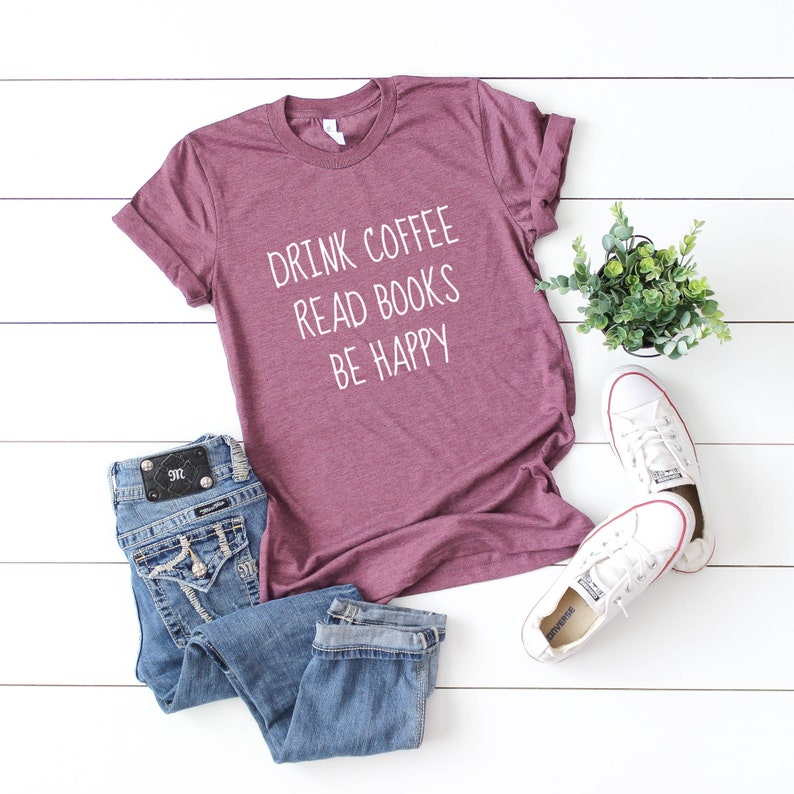 May include: A maroon t-shirt with the text "Drink Coffee Read Books Be Happy" in white lettering. The shirt is laid flat on a white wooden surface with a pair of blue jeans and white sneakers.
