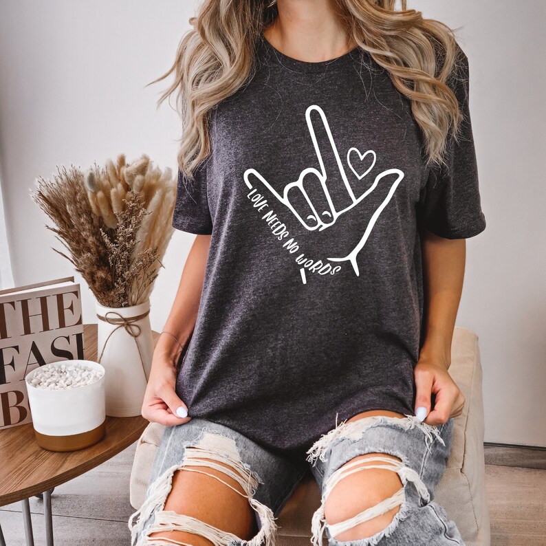 K&ouml;nnte beinhalten: Ein dunkelgraues T-Shirt mit einer wei&szlig;en Grafik einer Hand, die das Zeichen f&uuml;r 'Ich liebe dich' in amerikanischer Geb&auml;rdensprache macht. Die Grafik enth&auml;lt auch ein kleines Herz und den Text 'Liebe braucht keine Worte'.