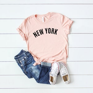 May include: A light pink t-shirt with the text "NEW YORK" in black lettering. The shirt is paired with blue jeans and white sneakers.