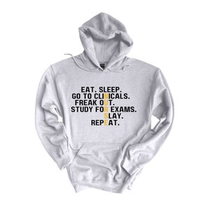 May include: A gray hoodie with the text "EAT. SLEEP. GO TO CLINICALS. FREAK OUT. STUDY FOR EXAMS. SLAY. REPEAT." in black with the word "NURSE" in yellow.