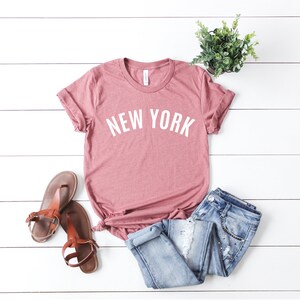 May include: A dusty rose t-shirt with the text "NEW YORK" in white letters. The shirt is laid out on a white wooden surface with a pair of brown sandals and blue jeans.