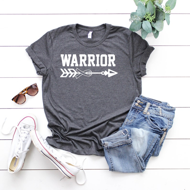 Puede incluir: Una camiseta gris oscuro con la palabra "WARRIOR" en texto blanco y una flecha con una cinta blanca en el centro. La camiseta est&aacute; colocada sobre una superficie de madera blanca con un par de zapatillas blancas, unos vaqueros azules y unas gafas de sol.