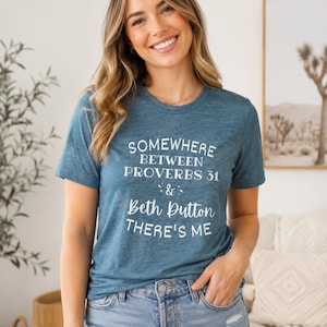 May include: A heathered teal short-sleeve t-shirt featuring white screen-printed text that reads: SOMEWHERE BETWEEN PROVERBS 31 & BETH DUTTON THERE'S ME. The shirt has a crew neckline and is paired with light blue denim jeans with a distressed detail.