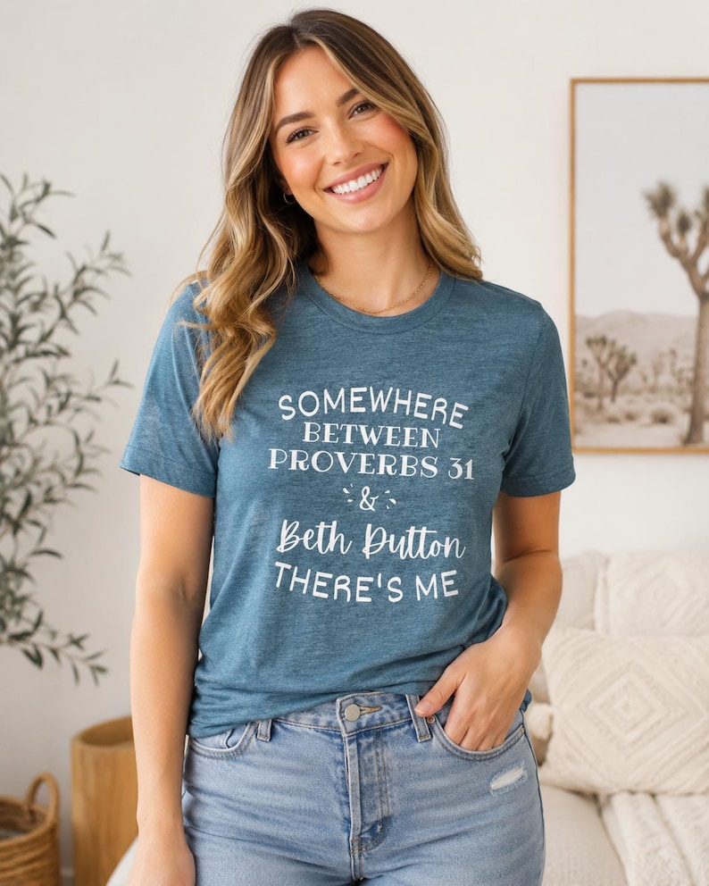 May include: A heathered teal short-sleeve t-shirt featuring white screen-printed text that reads: SOMEWHERE BETWEEN PROVERBS 31 & BETH DUTTON THERE'S ME. The shirt has a crew neckline and is paired with light blue denim jeans with a distressed detail.