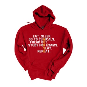 May include: Red hooded sweatshirt with the text "EAT. SLEEP. GO TO CLINICALS. FREAK OUT. STUDY FOR EXAMS. SLAY. REPEAT." in white with the word "NURSE" in yellow.