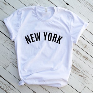 May include: White t-shirt with black text that reads "NEW YORK" in a curved arch.