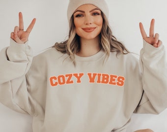 May include: A beige sweatshirt with the words "COZY VIBES" in orange block letters. The sweatshirt is worn with a beige beanie. The person is giving a peace sign with both hands.