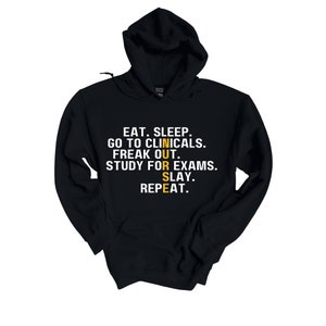 May include: Black hooded sweatshirt with white text that reads "EAT. SLEEP. GO TO CLINICALS. FREAK OUT. STUDY FOR EXAMS. SLAY. REPEAT." The word "NURSE" is spelled vertically in yellow.