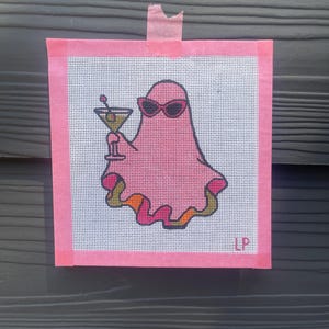 May include: A square needlepoint canvas featuring a pink ghost wearing dark sunglasses and holding a martini glass. The ghost has a wavy bottom edge with orange and green accents. The letters LP are printed in the bottom right corner of the canvas.
