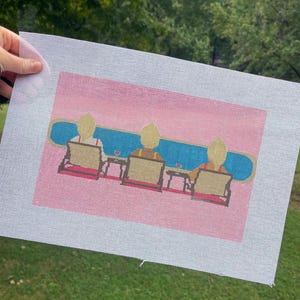 May include: A needlepoint canvas featuring a printed design of three figures with blonde hair sitting in lounge chairs on a pink background. The figures face a long blue horizontal shape representing water, with small side tables and glasses between them.