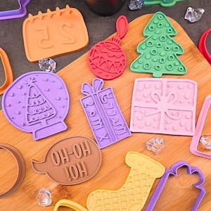 Peut inclure: Diverses formes de découpoirs à biscuits sur le thème de Noël, aux couleurs vives. On y trouve un sapin, une boule de Noël, un cadeau, une chaussette et une bulle de dialogue avec le texte "OH-OH-OH".