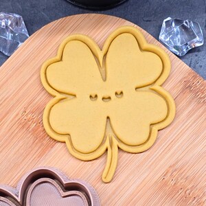 Op de afbeelding: Een gele klavervier koekjesvorm met een lachend gezicht, geplaatst op een houten snijplank. De klavervier wordt omringd door een koperkleurige koekjesvorm en twee heldere, facetgeslepen objecten.