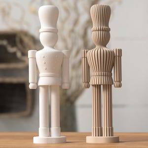 Op de afbeelding: Twee staande notenkraker-beeldjes. Het linkerfiguur is effen wit met een matte, getextureerde afwerking. Het rechterfiguur is lichtbruin en opgebouwd uit verticale latten, wat zorgt voor een geribbelde, gelaagde uitstraling met zichtbare kieren.