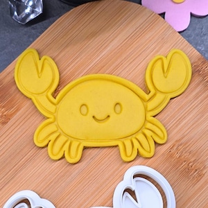 Può includere: Tagliapasta a forma di granchio giallo con un volto sorridente. Il tagliapasta è realizzato in plastica e presenta un design dettagliato. Il granchio è su un tagliere di legno, con un fiore e altri oggetti sullo sfondo.