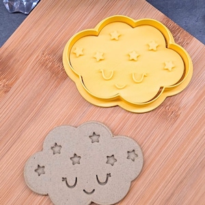Può includere: Un tagliabiscotti giallo a forma di nuvola con ritagli a forma di stella e una faccia sorridente. Un biscotto cotto a forma di nuvola con ritagli a forma di stella e una faccia sorridente è accanto al tagliabiscotti. Il biscotto e il tagliabiscotti sono su una superficie di legno.