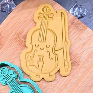 Pode incluir: Cortador de biscoitos castanho claro em forma de violino de desenho animado com arco. O violino tem um rosto sorridente e está em uma tábua de corte de madeira. Um cortador de biscoitos azul em forma de violino está no canto inferior esquerdo.