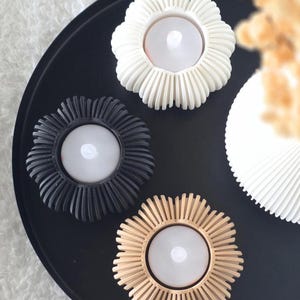 Può includere: Tre portacandele a forma di fiore nei colori bianco, nero e beige, ciascuno con un'apertura circolare centrale per una candela. I portacandele presentano un design esterno a coste simile a petali e sono disposti su un vassoio nero.