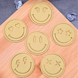 Pode incluir: Um conjunto de seis cortadores de biscoitos redondos com desenhos de rostos sorridentes. Cada cortador apresenta um design de olhos diferente, incluindo corações, estrelas e raios. Os cortadores são feitos de um material castanho claro e estão dispostos numa tábua de cortar de madeira.