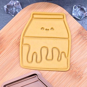 May include: A yellow cookie cutter shaped like a milk carton with a smiling face and dripping liquid design. The cutter is on a wooden board with a second cutter and decorative ice cubes.