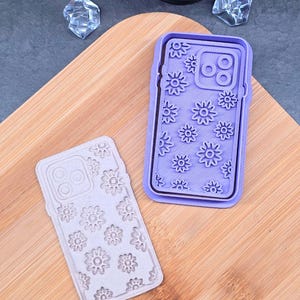 Peut inclure: Deux coques de téléphone avec un motif floral. L'une est violet clair, l'autre est beige clair. Les deux coques ont un motif de fleurs et une découpe pour l'appareil photo. Les coques sont sur une surface en bois.