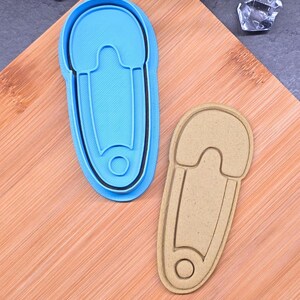 Peut inclure: Deux emporte-pièces en forme d'épingle à nourrice. L'un est bleu clair avec un contour noir, et l'autre est beige. Les deux sont en plastique et reposent sur une surface en bois. Les emporte-pièces sont conçus pour créer des biscuits en forme d'épingle à nourrice.