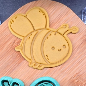 Può includere: Un tagliabiscotti a forma di ape gialla con un volto sorridente, ali e corpo a strisce. Il tagliabiscotti è in plastica e si trova su un tagliere di legno. L'ape ha un design carino e da cartone animato.