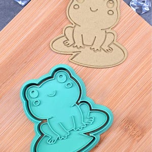May include: Two frog-shaped cookie cutters. One is tan, showing the outline of a frog on a lily pad. The other is teal, with a frog and lily pad design. Both cutters have a smiling frog face.