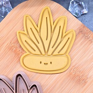 May include: A yellow cookie cutter in the shape of a potted succulent plant with a smiling face. The cutter is on a wooden cutting board, with a second, unformed cutter below.