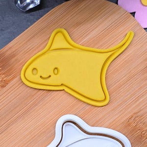 May include: A yellow cookie cutter in the shape of a stingray with a smiling face. The cutter is on a wooden cutting board. A white stingray cookie cutter is below the yellow one.