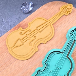 May include: Two violin-shaped cookie cutters, one in yellow and the other in teal, sit on a wooden cutting board. The yellow cutter is a solid shape, while the teal one has a layered design. A clear, faceted object is in the background.
