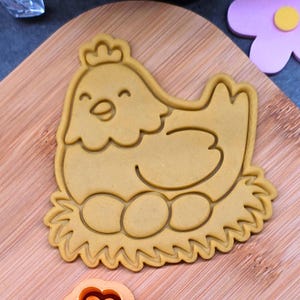 Può includere: Tagliapasta marrone chiaro a forma di gallina su un nido con tre uova. La gallina ha una faccia sorridente e una corona. Il tagliapasta è su un tagliere di legno.