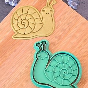 May include: Two snail-shaped cookie cutters. One is tan, the other is teal. Both have a spiral shell design and a smiling face. The cutters are on a wooden chopping board.