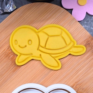 Könnte beinhalten: Ein gelber Ausstecher in Schildkrötenform mit einem lächelnden Gesicht und einem segmentierten Panzer. Der Ausstecher ist aus Kunststoff und liegt auf einem Holzschneidebrett. Eine rosa Blume und ein weiterer Ausstecher sind ebenfalls zu sehen.