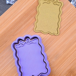 May include: Two cookie cutters in the shape of a tag with a bow at the top. One is light purple, the other is tan. Both have a wavy border. The cutters are on a wooden surface.