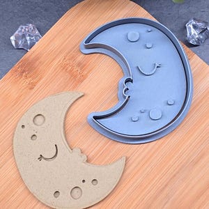 Pode incluir: Um cortador de biscoitos de plástico azul claro em forma de lua crescente ao lado de um biscoito bege em forma de lua crescente sorridente. O cortador de biscoitos tem um rosto sorridente e vários círculos pequenos. O biscoito tem um rosto sorridente e vários círculos pequenos.