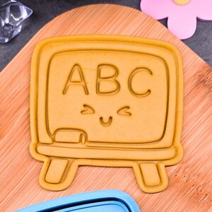 Könnte beinhalten: Gelber Ausstecher in Form einer Tafel mit den Buchstaben "ABC" und einem lächelnden Gesicht. Er liegt auf einem Holzbrett, mit einem blauen Ausstecher und einer rosa Blume im Hintergrund. Ideal zum Backen.