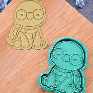 Op de afbeelding: Twee koekjessnijders in de vorm van een schildpad. Eén is beige, de andere is turquoise. Beide snijders hebben een lachende schildpad met een bril. De snijders liggen op een houten snijplank.
