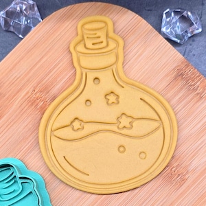 May include: A light brown cookie cutter in the shape of a potion bottle with a cork top. The bottle has a wavy line and star shapes inside. The cutter is on a wooden cutting board with clear, faceted decorative objects.